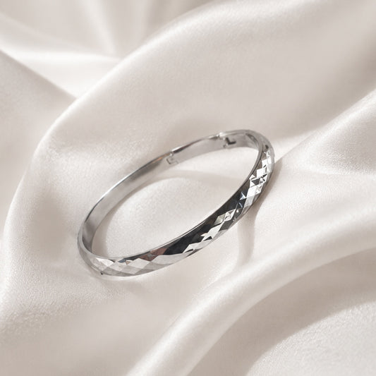 Faceted polished silver bangle bracelet displayed on ivory silk fabric in luxury editorial styling.