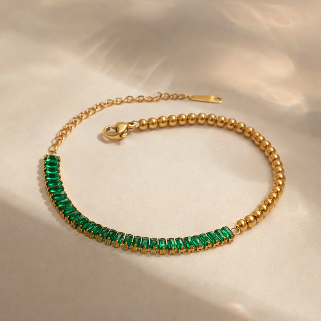 Gold bracelet featuring emerald baguette stones and polished gold bead detailing on white background.