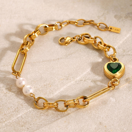 Gold charm link bracelet with emerald heart charm and pearl accents from OROR Parure on neutral stone background.