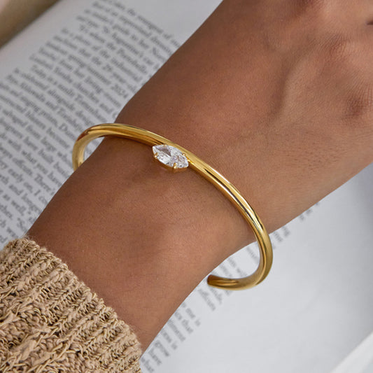 Gold bracelet with a clear gemstone on a wrist, with a blurred book in the background