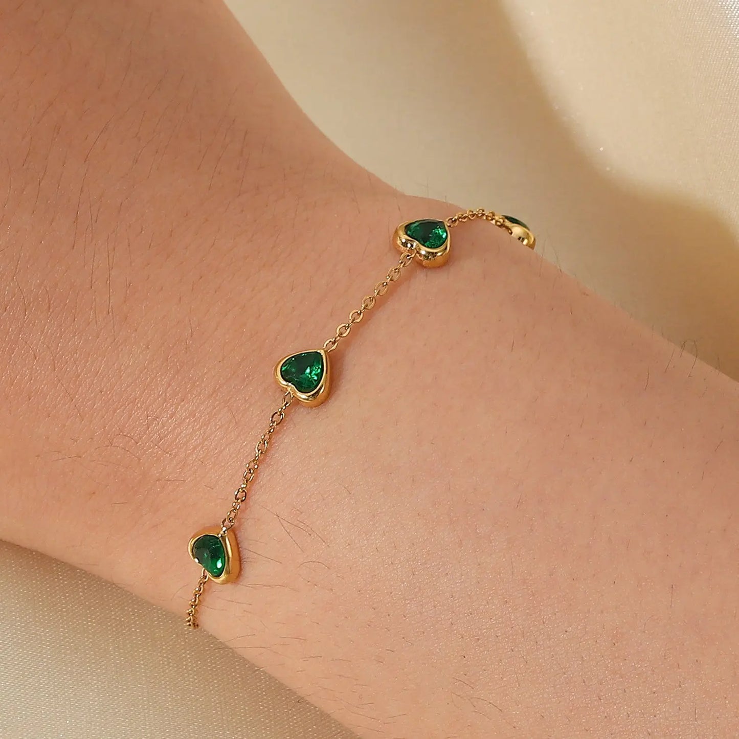 Gold bracelet with heart-shaped green gemstones on a wrist against a beige background