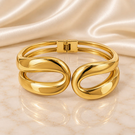 A polished gold-tone cuff bracelet with a double-band twisted design, displayed on a light marble surface with a draped satin fabric background.