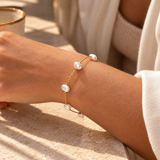 A delicate gold chain bracelet with four white baroque pearls, shown worn on a person's wrist in warm natural sunlight.