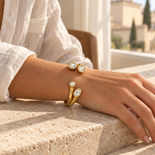 A close-up of a person's hand and forearm resting on a stone ledge, wearing two gold wrap-style bracelets with oval and round clear gemstones. The background shows a softly blurred outdoor architectural setting with warm sunlight.