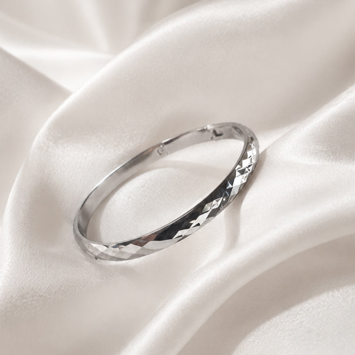 Faceted polished silver bangle bracelet displayed on ivory silk fabric in luxury editorial styling.