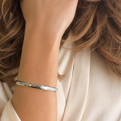 Faceted polished silver bangle bracelet displayed on ivory silk fabric in luxury editorial styling.