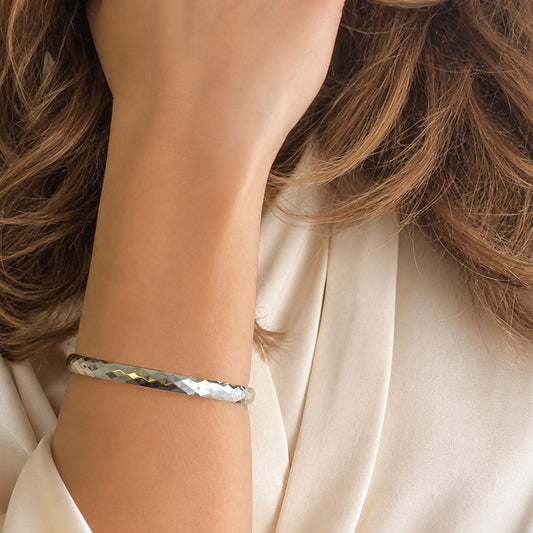 Faceted polished silver bangle bracelet displayed on ivory silk fabric in luxury editorial styling.