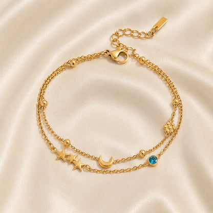 Double-chain gold bracelet featuring stars, a crescent moon, gold beads, and a blue crystal displayed on an ivory satin background.