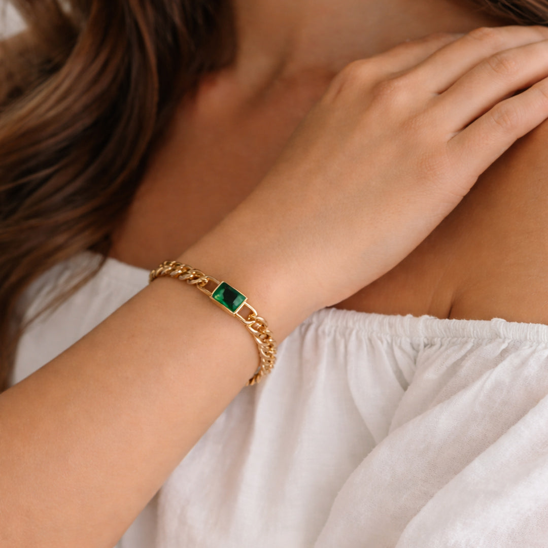Gold curb chain bracelet with emerald-cut green stone worn on sun-kissed wrist in white outfit