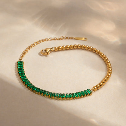 Gold bracelet featuring emerald baguette stones and polished gold bead detailing on white background.