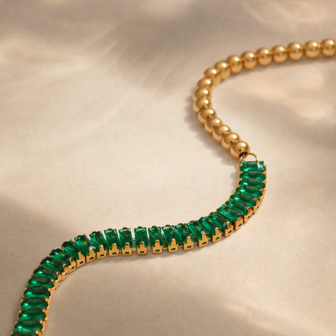Gold bracelet featuring emerald baguette stones and polished gold bead detailing on white background.