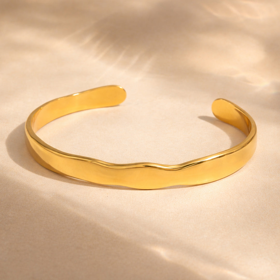 Polished sculpted open gold cuff bracelet with fluid curved silhouette on warm ivory luxury background.