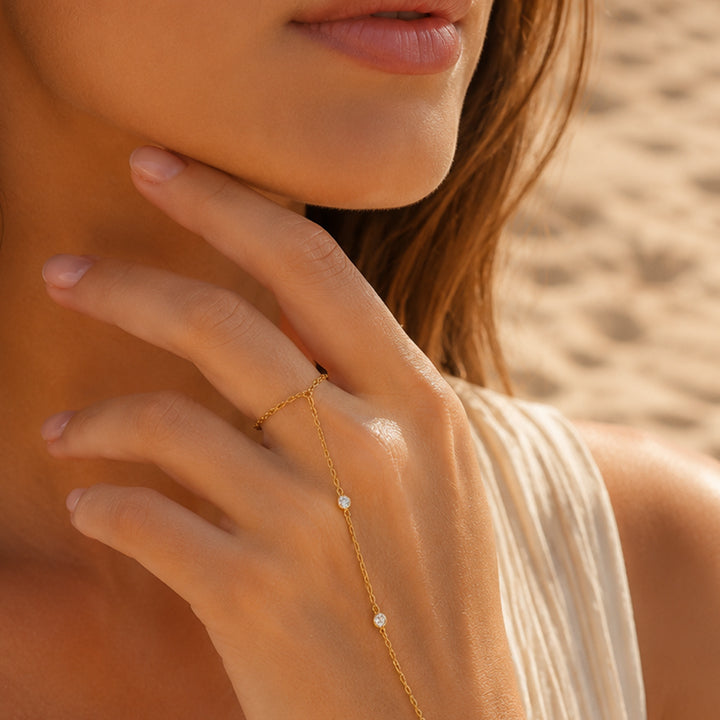 Close-up of a woman's hand wearing a delicate gold necklace with small stones, against a blurred natural background.