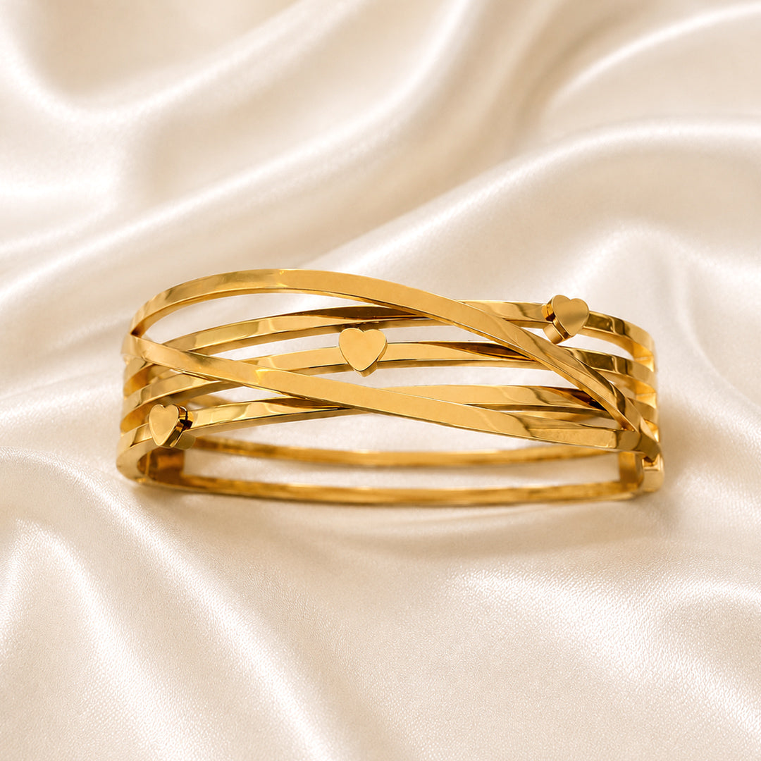 Amora Heart Bangle featuring intertwined gold bands with three polished heart accents on an ivory satin background.