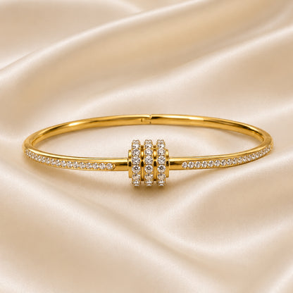 Slim 18K gold PVD bangle with a triple pavé ring centerpiece and cubic zirconia accents displayed on an ivory satin background.
