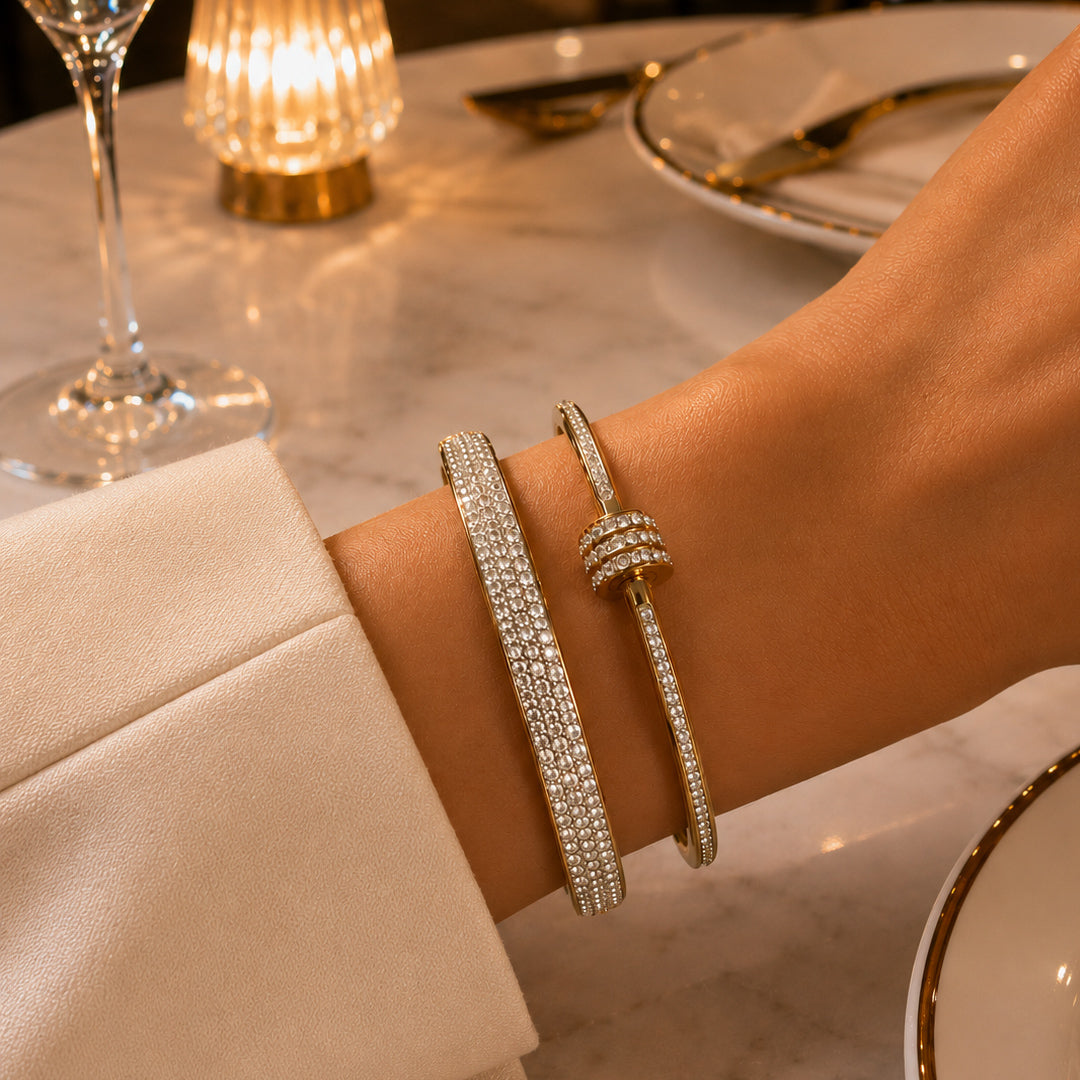 Close-up of a wrist wearing two gold bracelets with a blurred dinner setting in the background