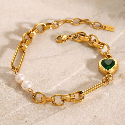 Gold charm link bracelet with emerald heart charm and pearl accents from OROR Parure on neutral stone background.