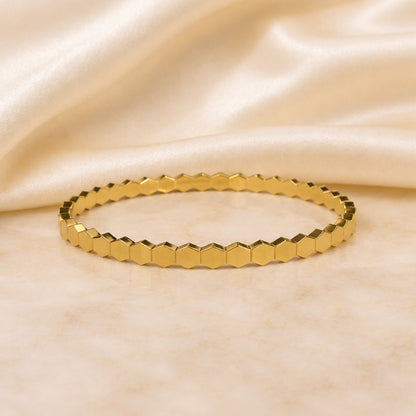 Faceted geometric gold bangle with hexagonal detailing displayed on an ivory satin and marble background.