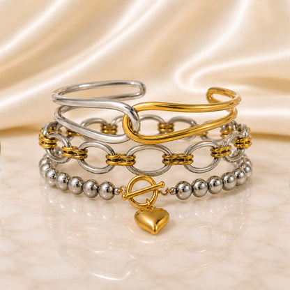 Three-piece mixed-metal bracelet stack featuring a sculptural two-tone cuff, interlocking chain bracelet and heart charm beaded bracelet displayed on ivory satin and marble.