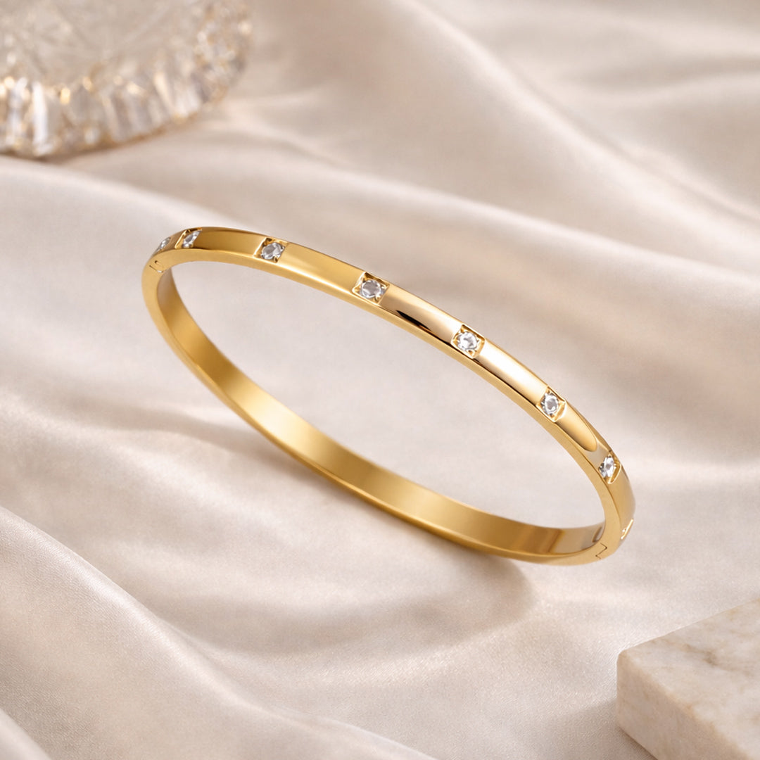 Slim gold bangle bracelet with bezel-set clear zircon stones on an ivory satin background