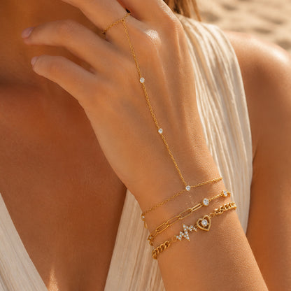 A close-up lifestyle shot of a model's hand and wrist wearing a gold chain hand bracelet with small white stones, layered with three gold bracelets on the wrist.