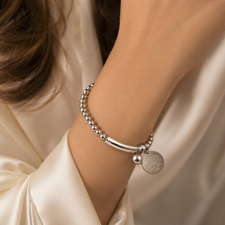Silver bracelet with a round tag on a person's wrist against a beige fabric background