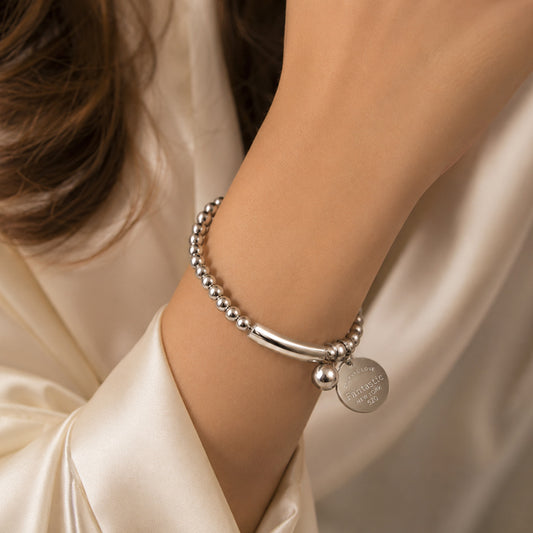 Silver bracelet with a round tag on a person's wrist against a beige fabric background
