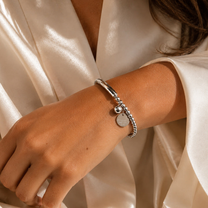 Luxury silver sphere charm bracelet with polished bead design and sculptural curved centerpiece worn on sun-kissed Mediterranean skin against ivory satin styling.