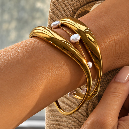 A close-up of a person's wrist wearing a stack of gold bangles, including two thick polished gold cuffs and delicate gold chains with white baroque pearls, set against a neutral textured background.