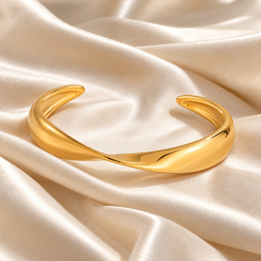 A polished gold cuff bracelet with a smooth, flowing organic curve, displayed on draped cream-colored satin fabric.