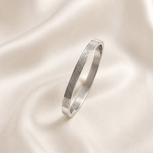 Aurelios Stainless Steel Greca Bangle featuring engraved geometric Greca detailing displayed on ivory satin.