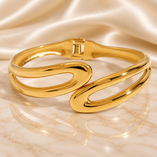A polished gold-tone cuff bracelet with an open, flowing double-loop design, displayed on a reflective marble surface with draped satin fabric in the background.