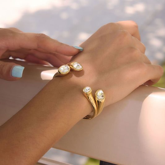 Gold wrap-style rings with clear oval gemstones worn on a person's wrist and hand, photographed outdoors with a softly blurred light background.