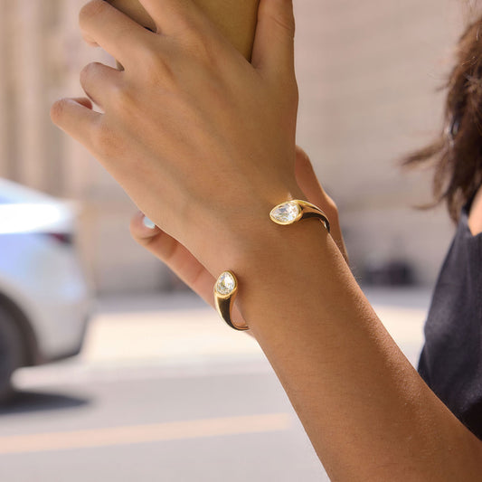 A close-up lifestyle shot of a person's wrist wearing a gold cuff bracelet with a marquise-cut clear gemstone. The background is a softly blurred outdoor street scene with a light-colored building and a white car.