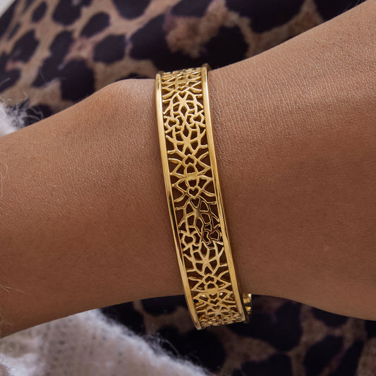 A gold-toned metal bangle bracelet with an intricate filigree cutout pattern, shown worn on a person's wrist.