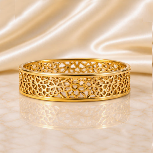 A gold bangle bracelet with an intricate openwork floral pattern, resting on a reflective marble surface with a draped satin fabric background.