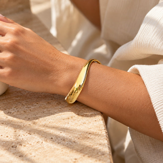 A polished gold bangle bracelet worn on a person's wrist, shown in a close-up lifestyle shot with warm sunlight and a textured stone surface.