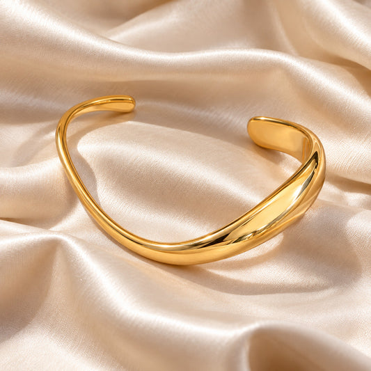 A polished gold-toned open cuff bracelet displayed on draped champagne satin fabric.