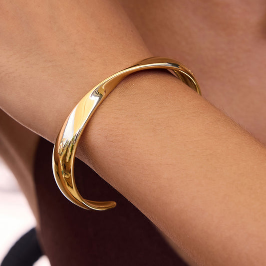 A polished gold-toned metal cuff bracelet worn on a person's wrist, shown in a close-up shot against skin tones.