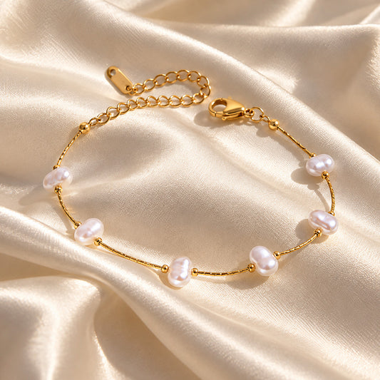A delicate gold chain bracelet with irregular white freshwater pearls, displayed on a draped cream satin fabric background.
