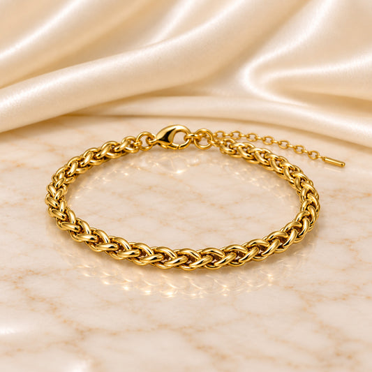 A gold-toned braided chain bracelet with a lobster clasp and extender chain, displayed on a light marble surface with draped satin fabric in the background.