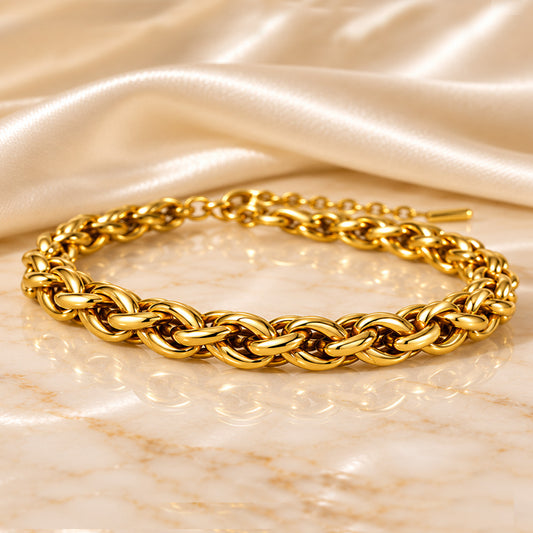 A gold-toned braided chain bracelet with a polished finish and a small gold clasp, displayed on a light beige marble surface with draped satin fabric in the background.