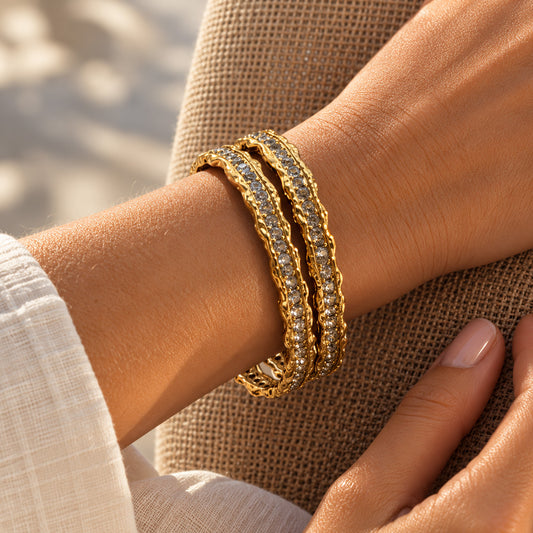 A close-up of a person's wrist wearing three stacked gold bangles, each set with small clear stones, against a neutral textured fabric background.