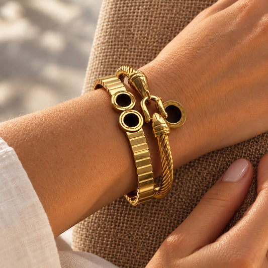 A close-up of a person's wrist wearing a gold-toned bracelet set with a flat link band, a rope-style bangle, and circular connector rings, shown against a neutral textured fabric background.