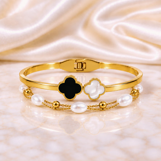 A gold-plated bangle bracelet featuring two clover motifs, one black and one white, accented by a delicate chain of white pearls and gold beads, displayed on a white marble surface with a cream satin background.
