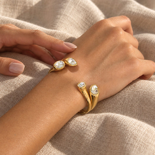 A close-up of a person's hand and wrist wearing two gold wrap-style rings with oval clear stones, resting on a beige linen fabric background.