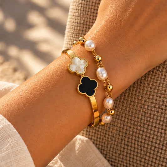 Two gold-toned bracelets displayed on a cream fabric pillow. One bracelet features a large black four-leaf clover charm with a white center, and the other is a pearl-bead chain with gold ball accents.