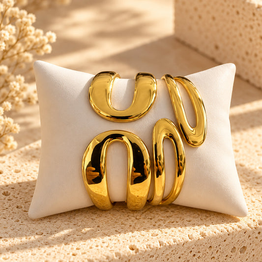 Four polished gold abstract sculptural wall art pieces arranged on a white rectangular pillow, set on a beige textured stone surface with dried floral decor in the background.